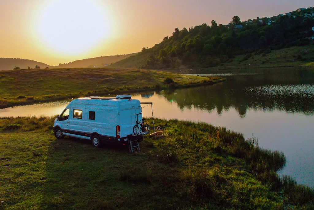 White camper van parked on grassy lakeshore at sunset, with calm water reflecting nearby hills and trees, and a bicycle mounted on the back.