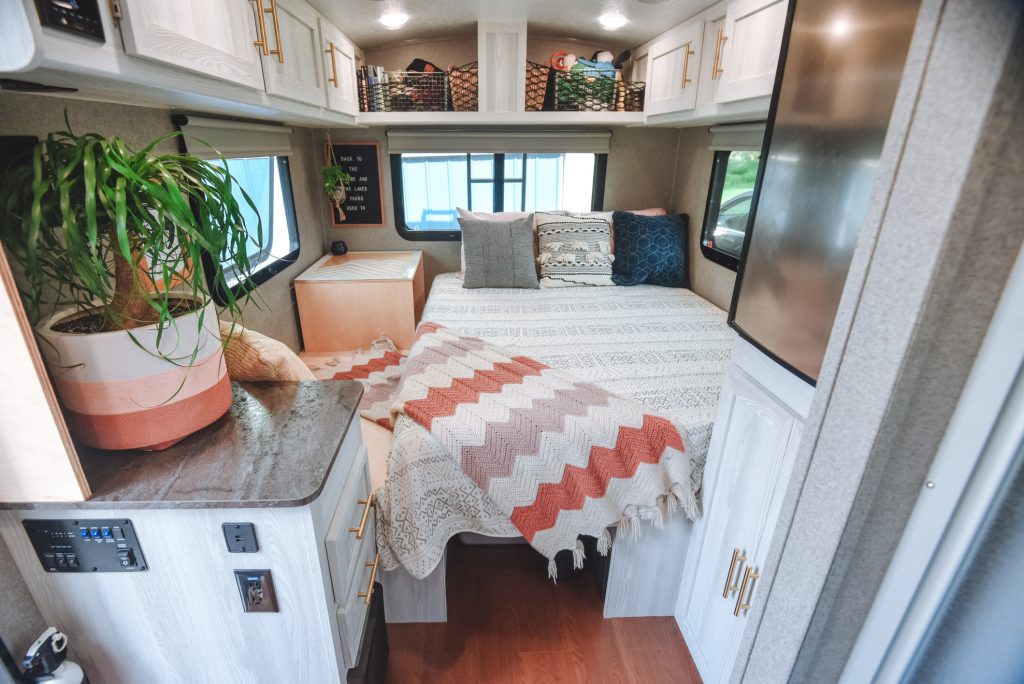 Cozy RV bedroom interior with a neatly made bed, patterned throw blanket and pillows, overhead cabinets, small nightstand, windows on three sides, and a potted plant on a nearby counter.
